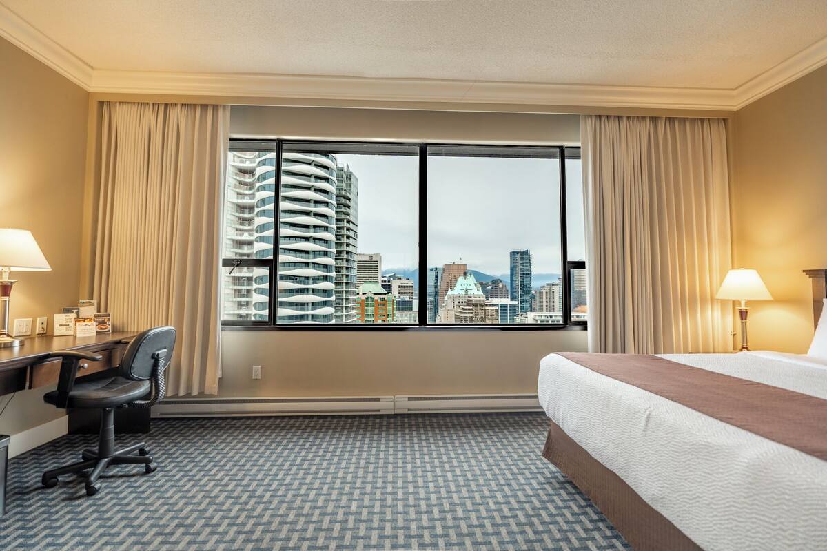 A hotel room with a large window, a bed, a desk, and a chair.