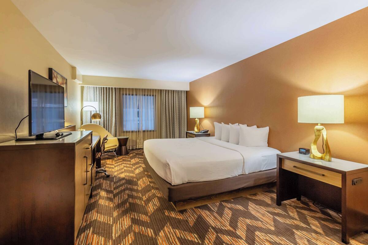 A hotel room with a large bed, a desk with a computer, a chair, and two bedside tables with lamps.