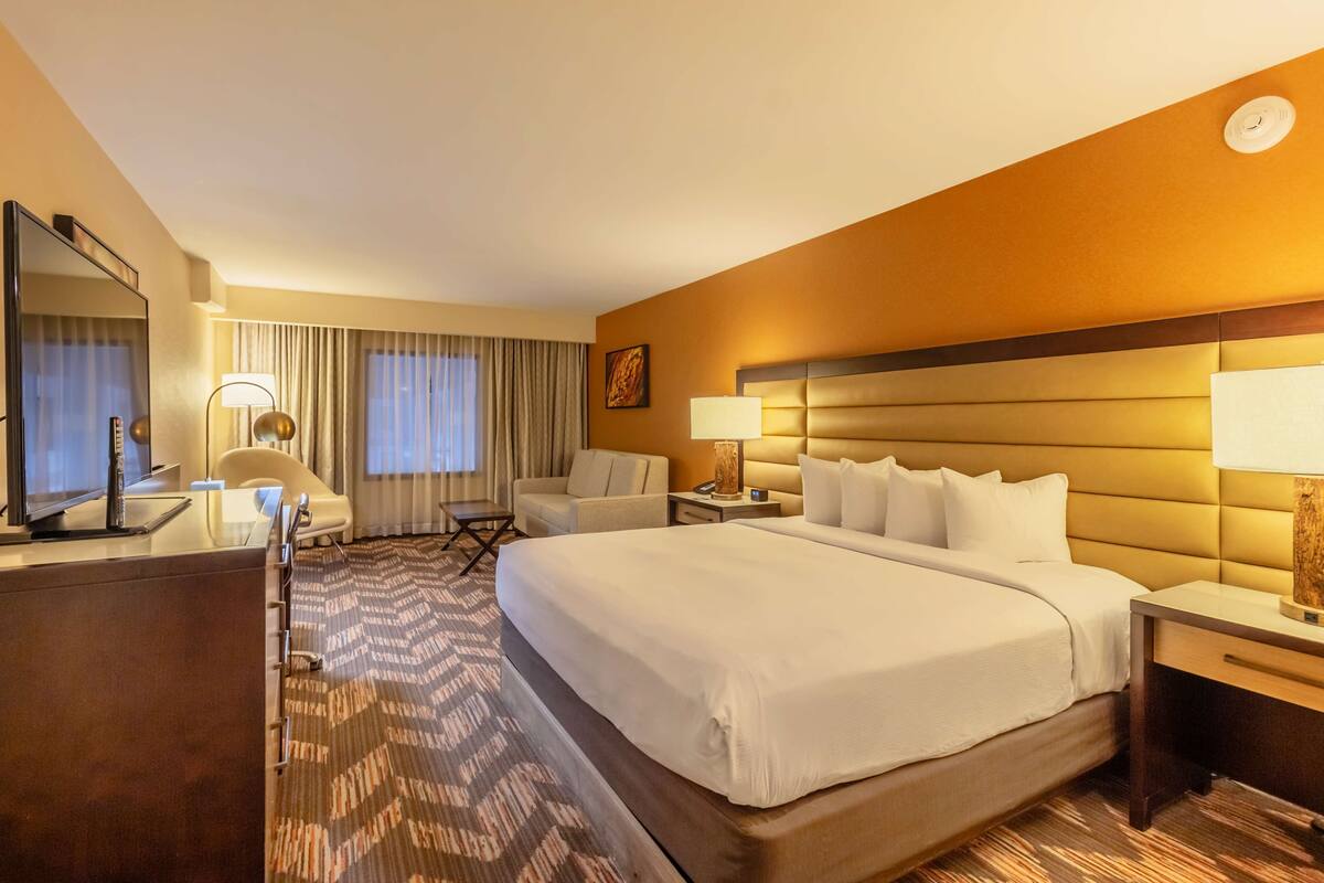A hotel room with a large bed, a desk with a computer, a chair, a lamp, and a window with curtains.