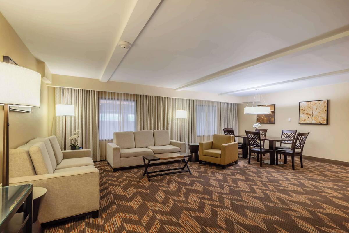 A hotel room with a sofa, armchair, dining table with chairs, and a patterned carpet.