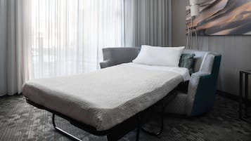 A hotel room with a bed, a grey armchair, a white pillow, a patterned carpet, and a floor lamp.