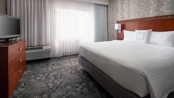 Habitación de hotel con una cama grande, un televisor de pantalla plana sobre un aparador de madera, una ventana con cortinas translúcidas y una alfombra estampada.