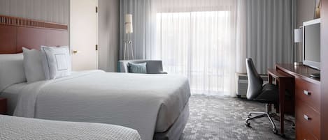 Habitación de hotel con dos camas, un escritorio, una silla, un televisor y un ventanal con cortinas.
