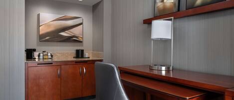 A modern hotel room with a wooden desk, a black leather chair, a large abstract painting, a white lamp, and a coffee maker on the counter.