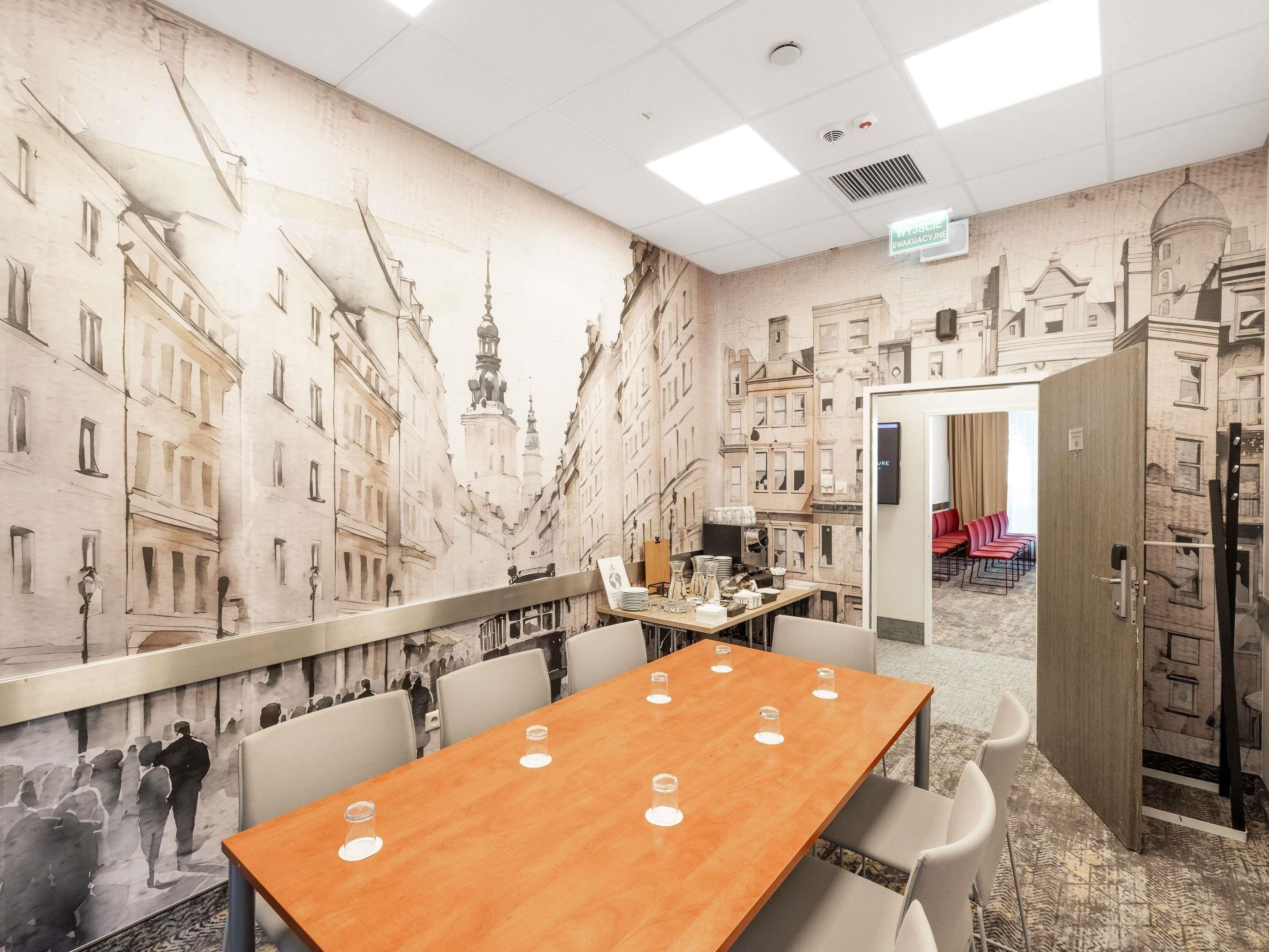 A conference room with a long table, chairs, a projector screen, and a mural of buildings on the wall.