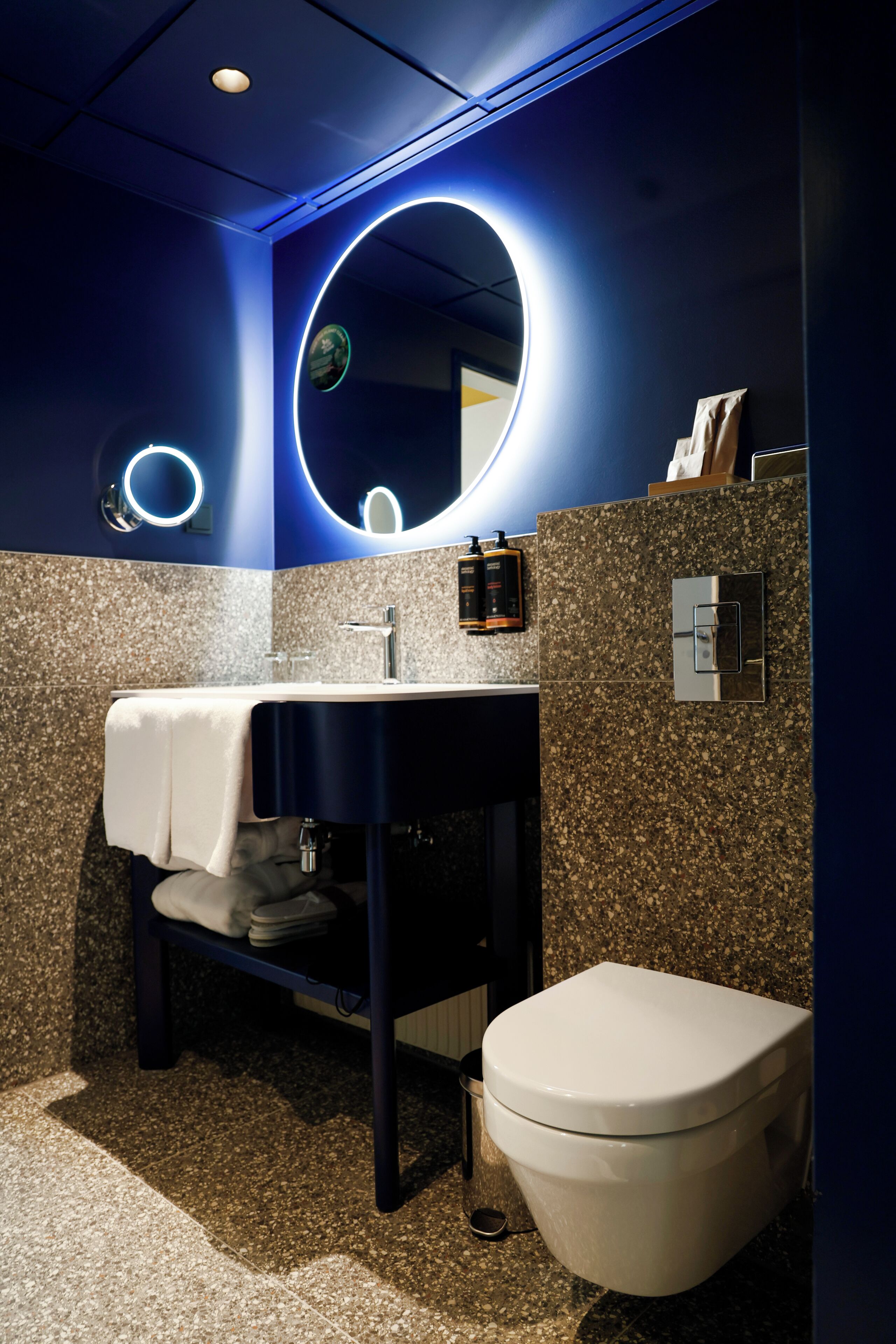 Un baño moderno con una pared azul oscuro, un lavabo con encimera de granito, un inodoro blanco e iluminación circular en un espejo.