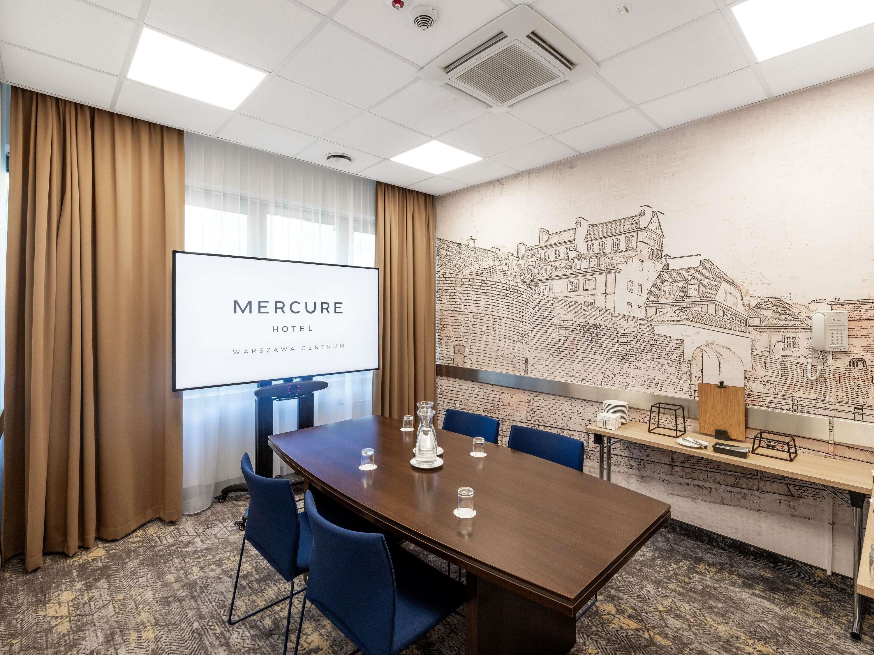 Un salón de conferencias con una gran mesa, sillas azules, una pantalla de proyector que muestra 'Mercure Hotel Warszawa Centrum' y un mural con un paisaje urbano en la pared.