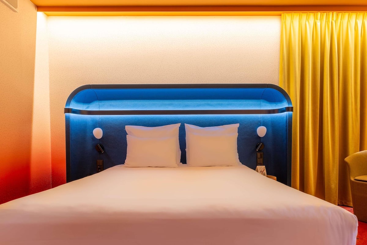 Una habitación de hotel con una cama grande, un cabecero con luces circulares y una cortina amarilla.
