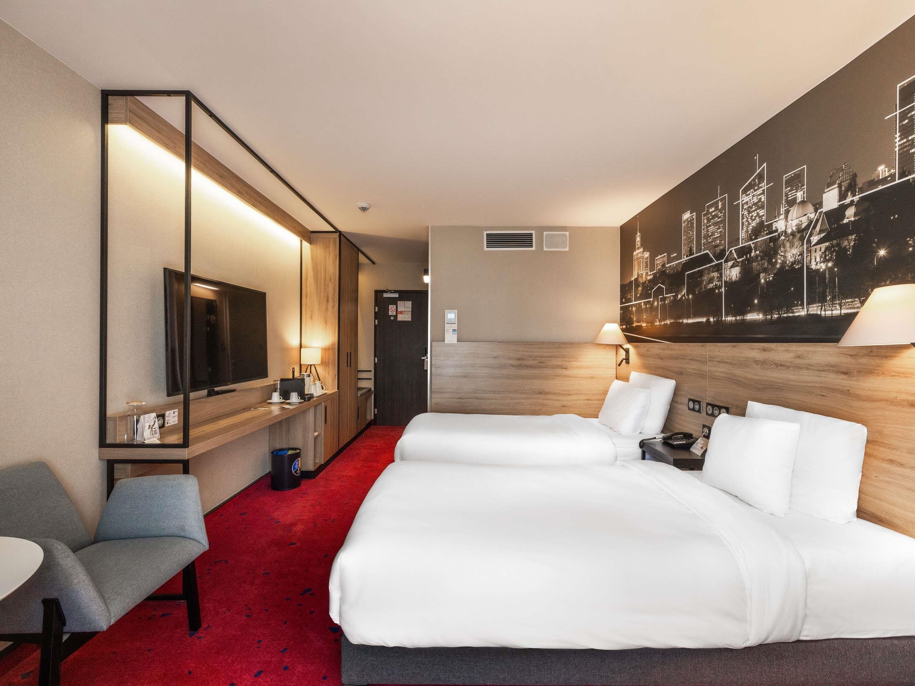 A hotel room with a large bed, a desk with a chair, a TV, and a cityscape mural on the wall.