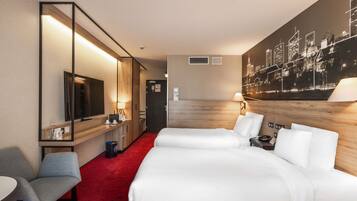 A hotel room with a large bed, a desk with a chair, a TV, and a cityscape mural on the wall.
