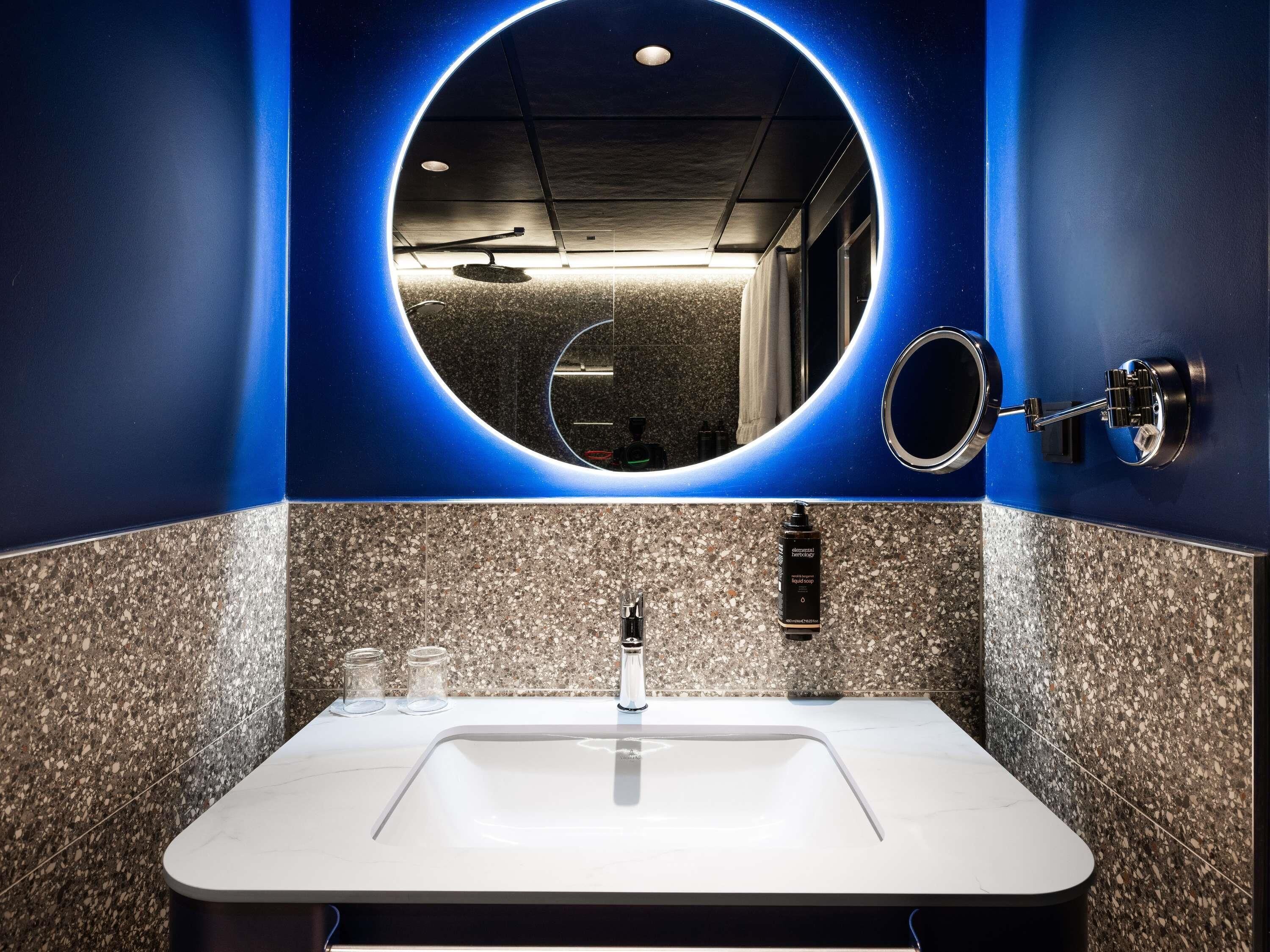 A bathroom with a white sink, granite tiles, a round mirror, and a blue accent wall.