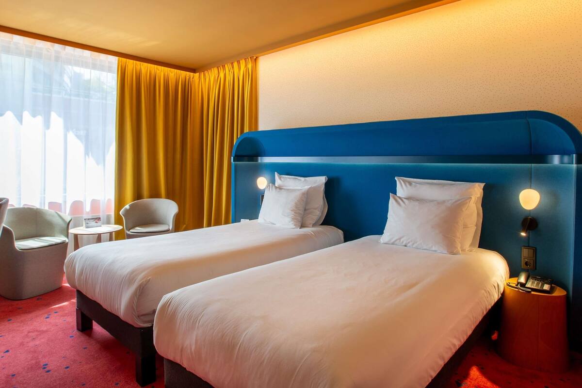 Habitación de hotel con dos camas, una mesita, una silla y una ventana con cortinas.