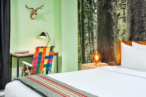 A hotel room with a bed, a desk with a lamp, a colorful chair, a deer head decoration, and a forest-themed wall.