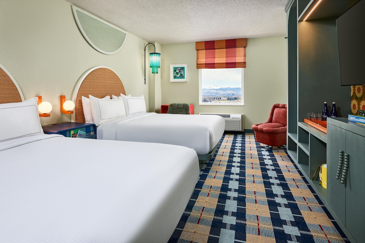 A hotel room with two beds, a red armchair, a minibar, and a view of a mountain landscape.