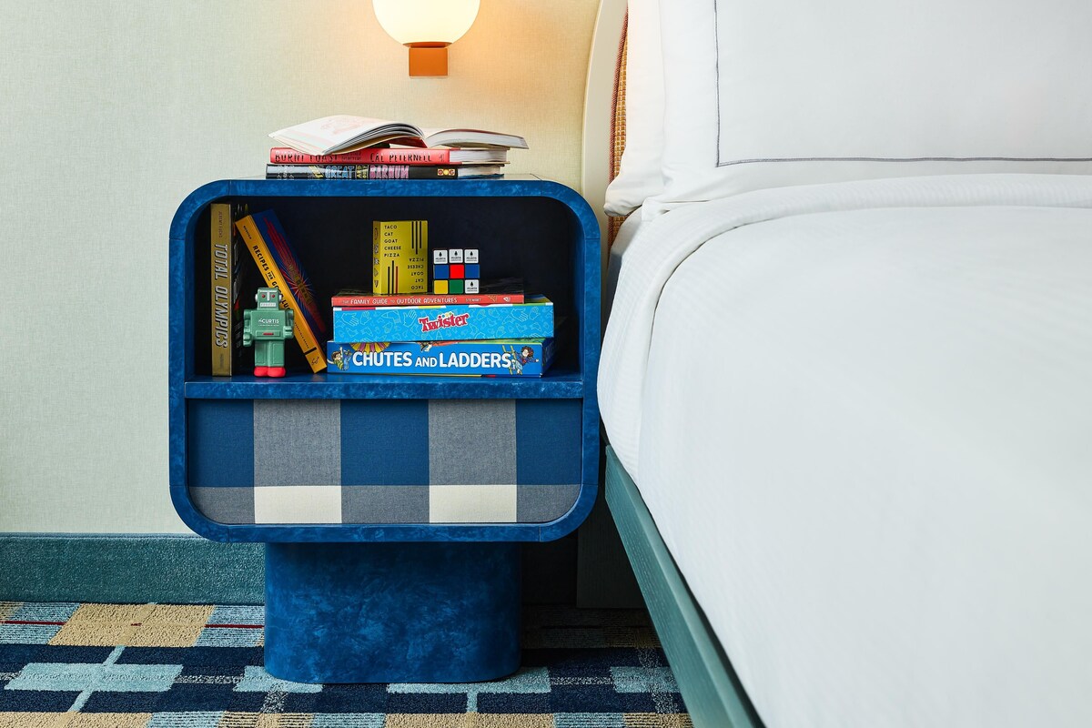 A neatly made bed with a white comforter and pillows, a blue bedside table with a lamp, and a small shelf with books and a toy.