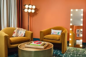 A room with two mustard-colored armchairs, a round coffee table with a book and a small box, a floor lamp, and a wall-mounted mirror with lights.