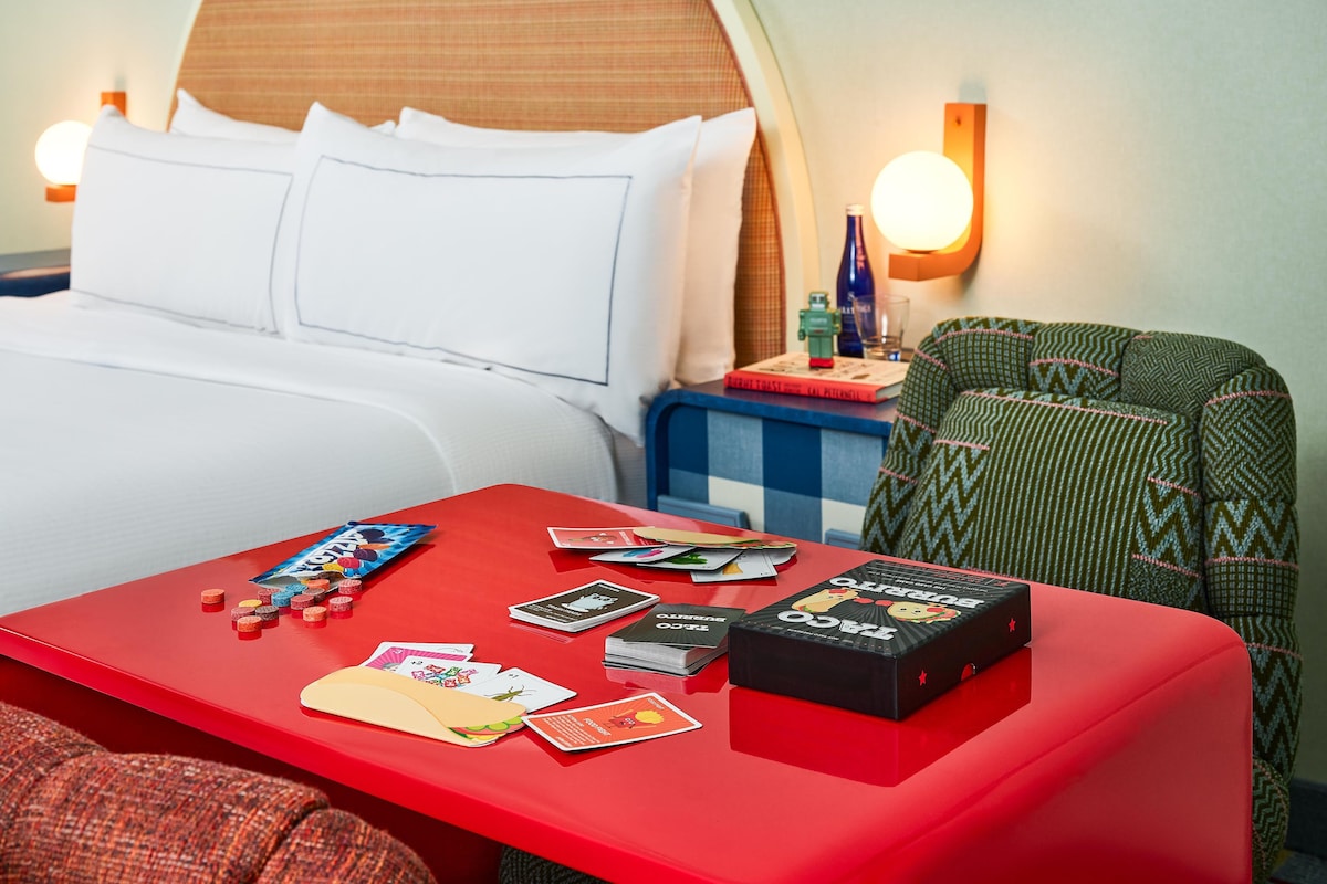 A hotel room with a bed, a red table with board games, and a green patterned armchair.