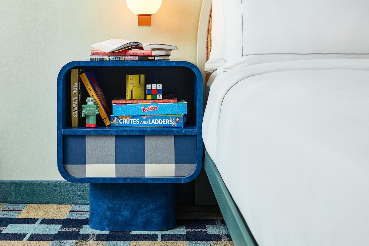 A neatly made bed with a white comforter and pillows, a small blue nightstand with a checkered pattern, and a wall-mounted light.