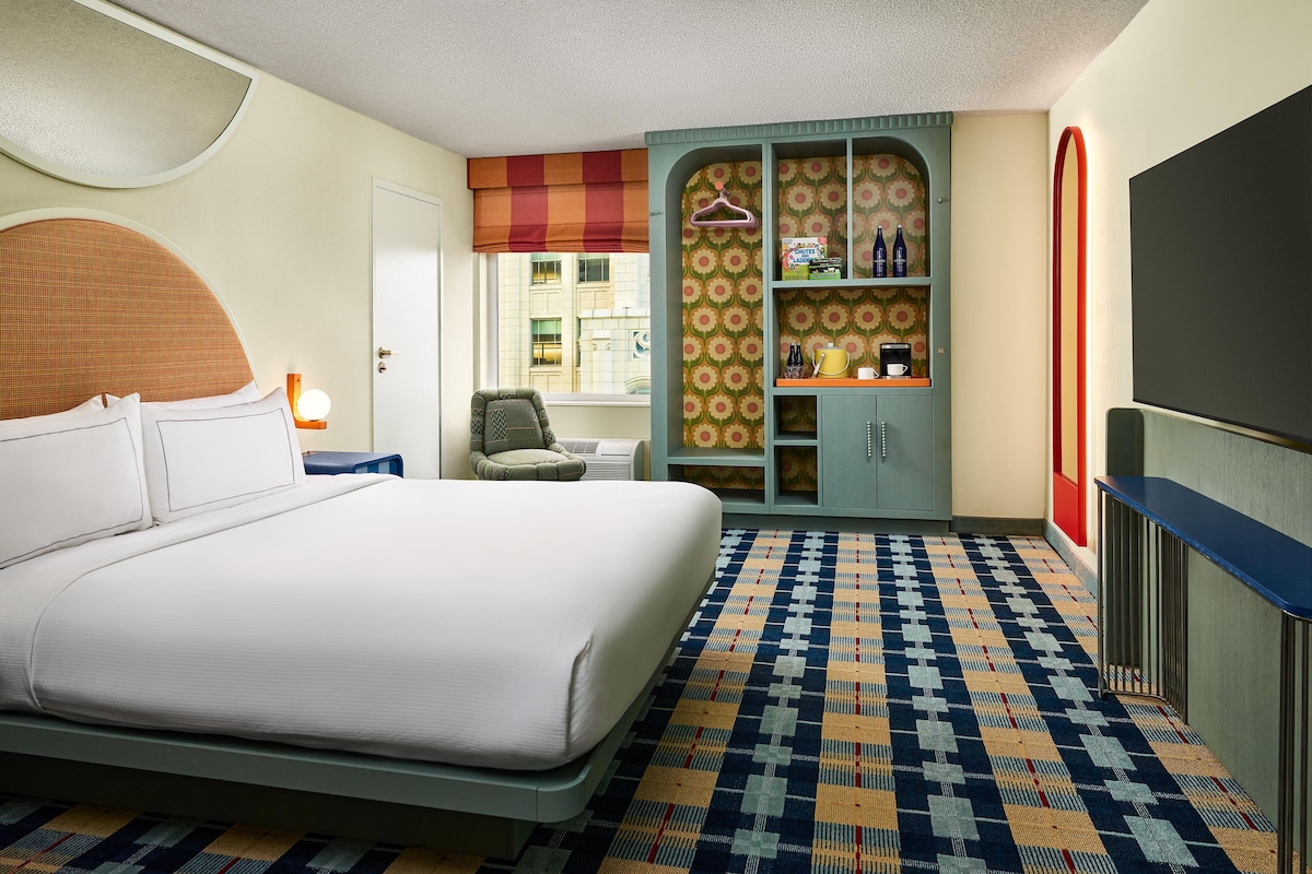 A hotel room with a large bed, a seating area with a chair, a wardrobe, and a television.