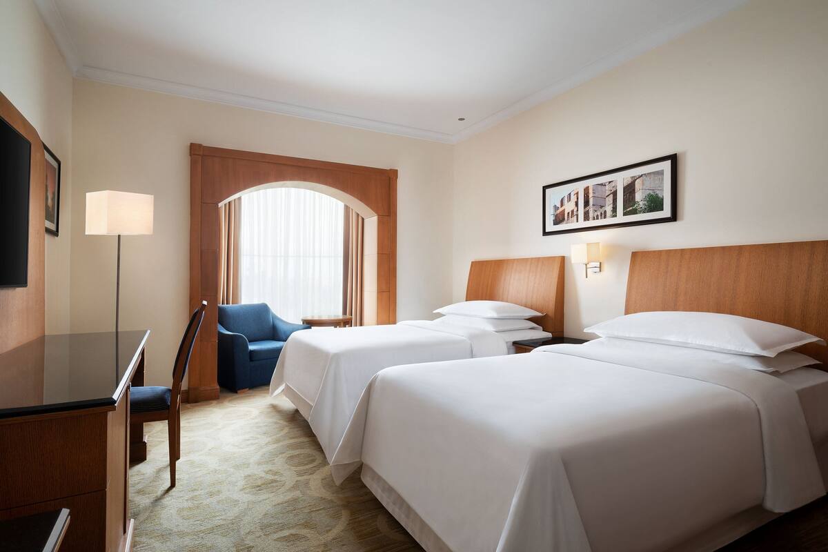 A hotel room with two beds, a desk, a chair, a TV, and a window with curtains.