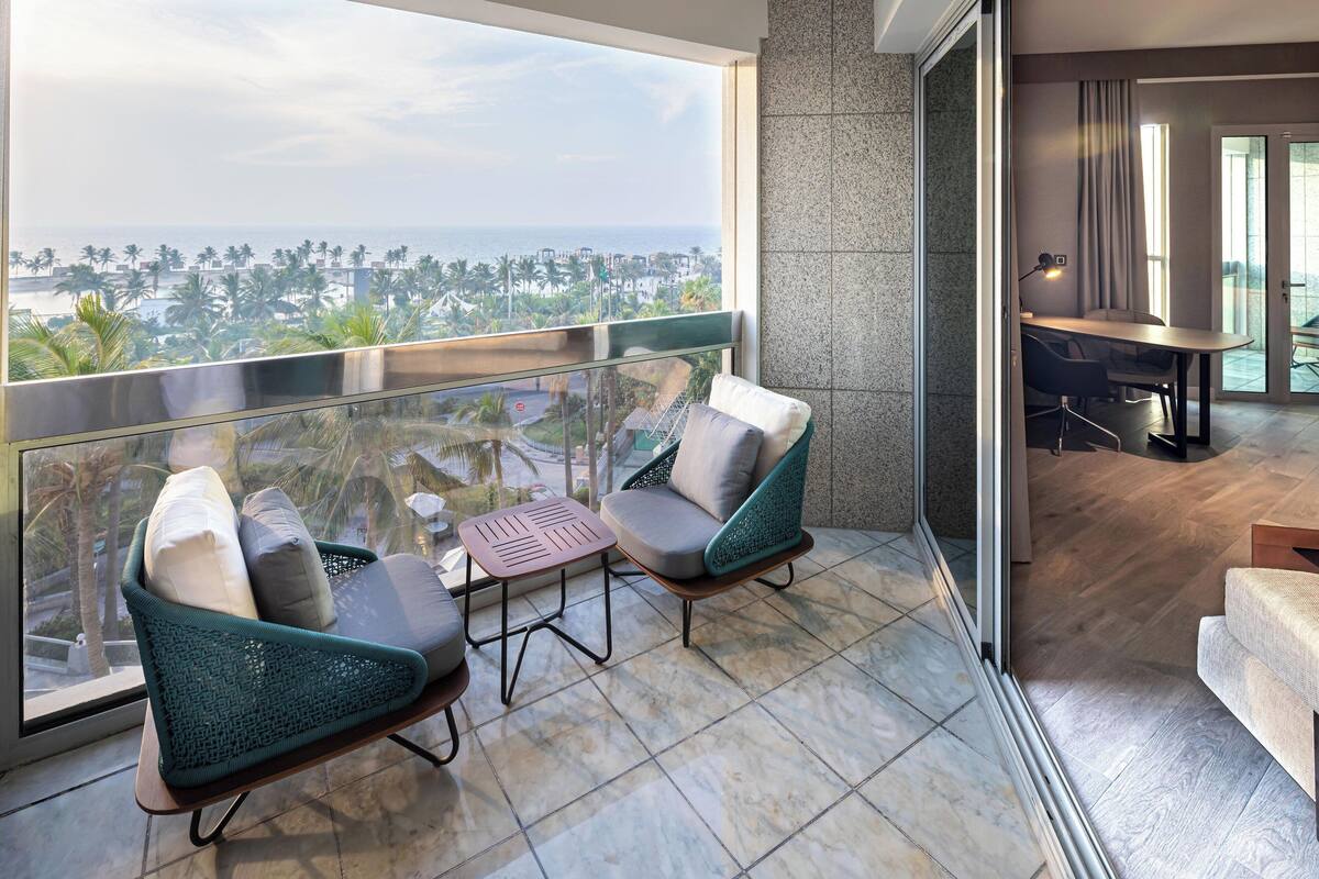 A balcony with two chairs and a small table, a view of the ocean, and a glimpse of a dining area.
