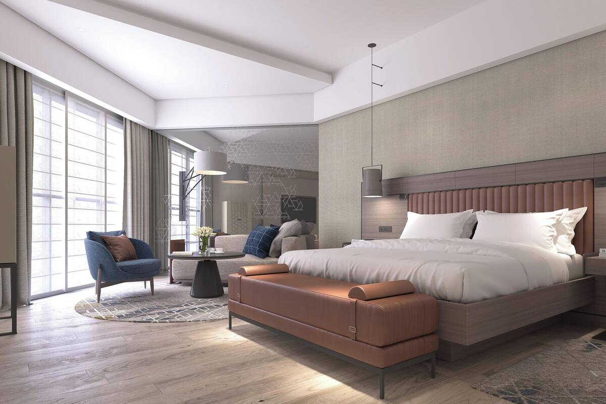 A modern hotel room with a large bed, a seating area with a sofa and armchair, a small table, and a large window providing natural light.