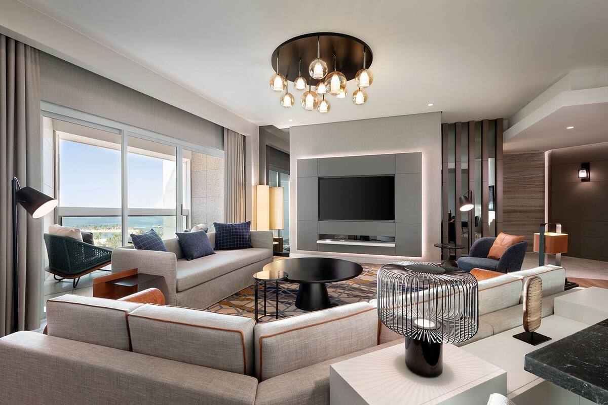 A modern hotel room with a large flat-screen TV, a sofa set, a coffee table, and a unique pendant light fixture.