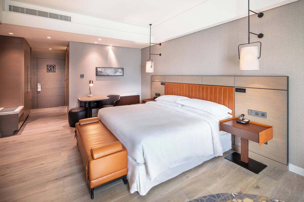 A modern hotel room with a large bed, a bedside table with a lamp, a small desk, and a bench.