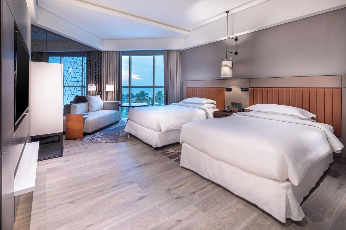 A hotel room with two beds, a sofa, a TV, and a large window with a city view.