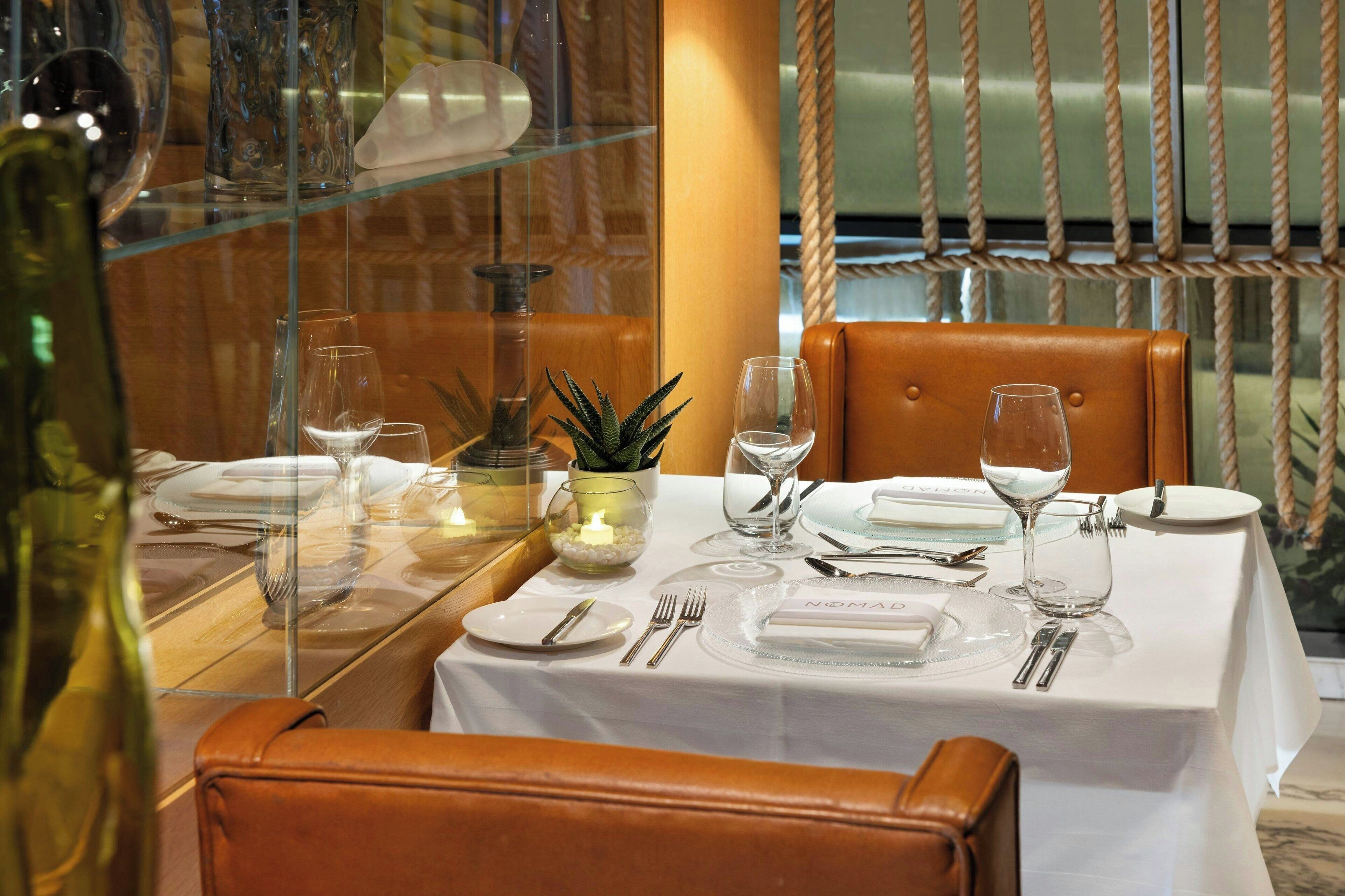 A dining table set with white tablecloth, plates, cutlery, and wine glasses, with a rope barrier and leather chairs.