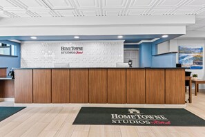 A reception area with a wooden reception desk, a digital payment terminal, and a reception desk mat with the property name. - HomeTowne Studios by Red Roof & Conference Center Cortland (Cortland)