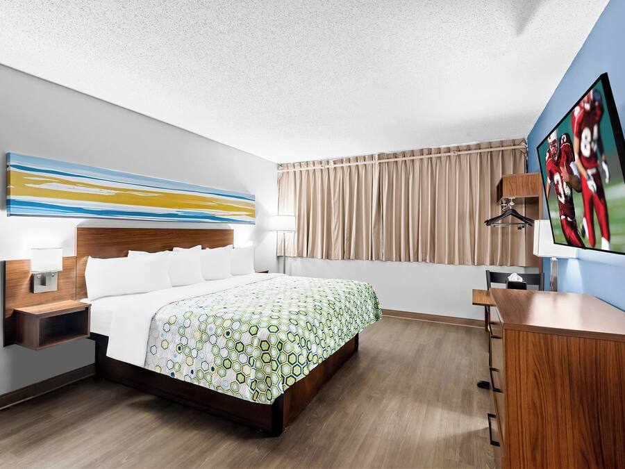 Habitación de hotel con cama, escritorio, televisor, una obra de arte en la pared y ventana con cortinas.