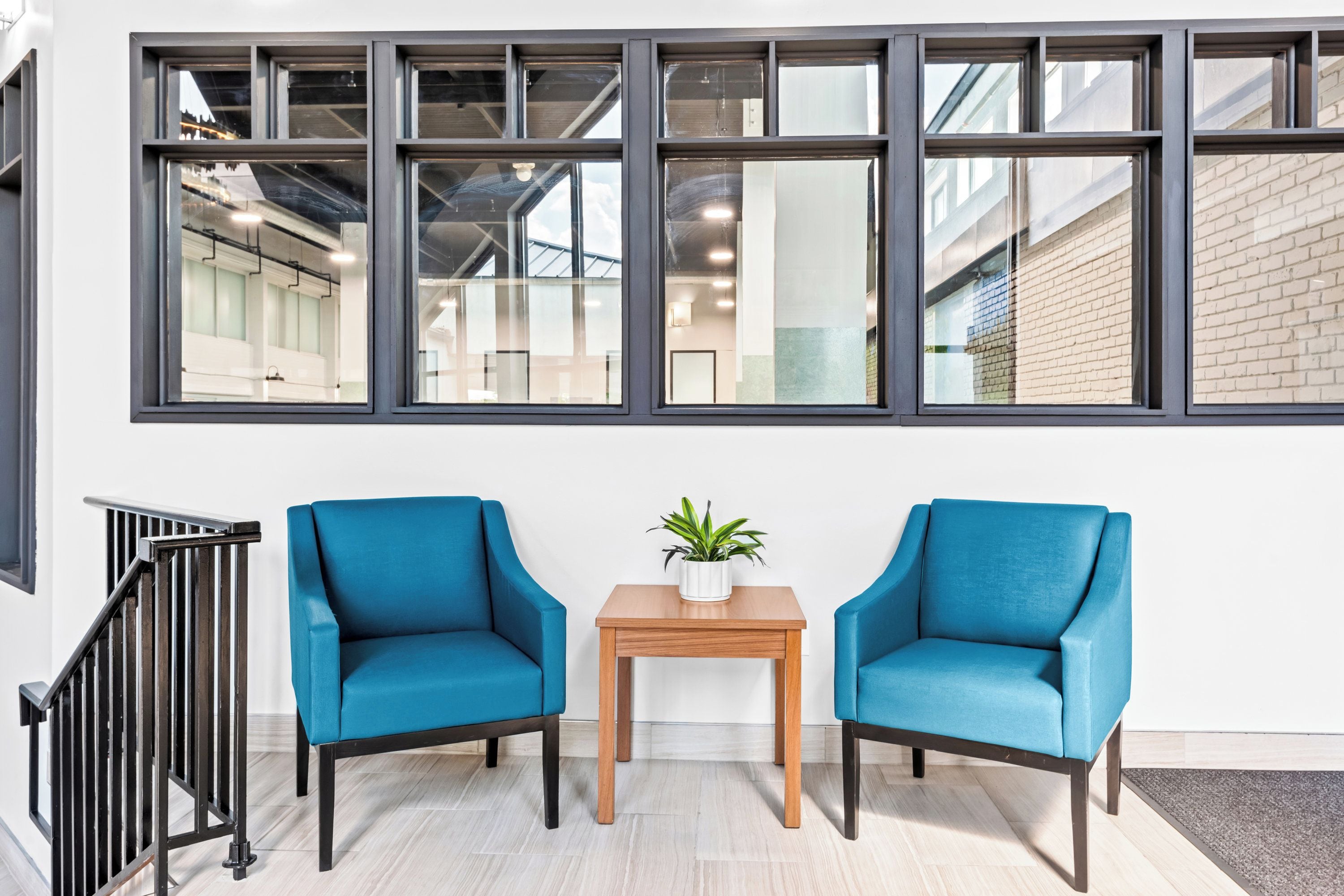 A modern interior with two blue armchairs, a small wooden table with a plant, and a large window with multiple panes.