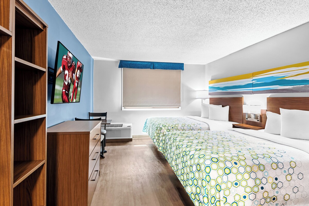 Een moderne hotelkamer met een houten bureau, een bed met een beddegoed met een geometrisch patroon, een groot raam met een blauwe zonwering en een aan de muur bevestigde televisie.