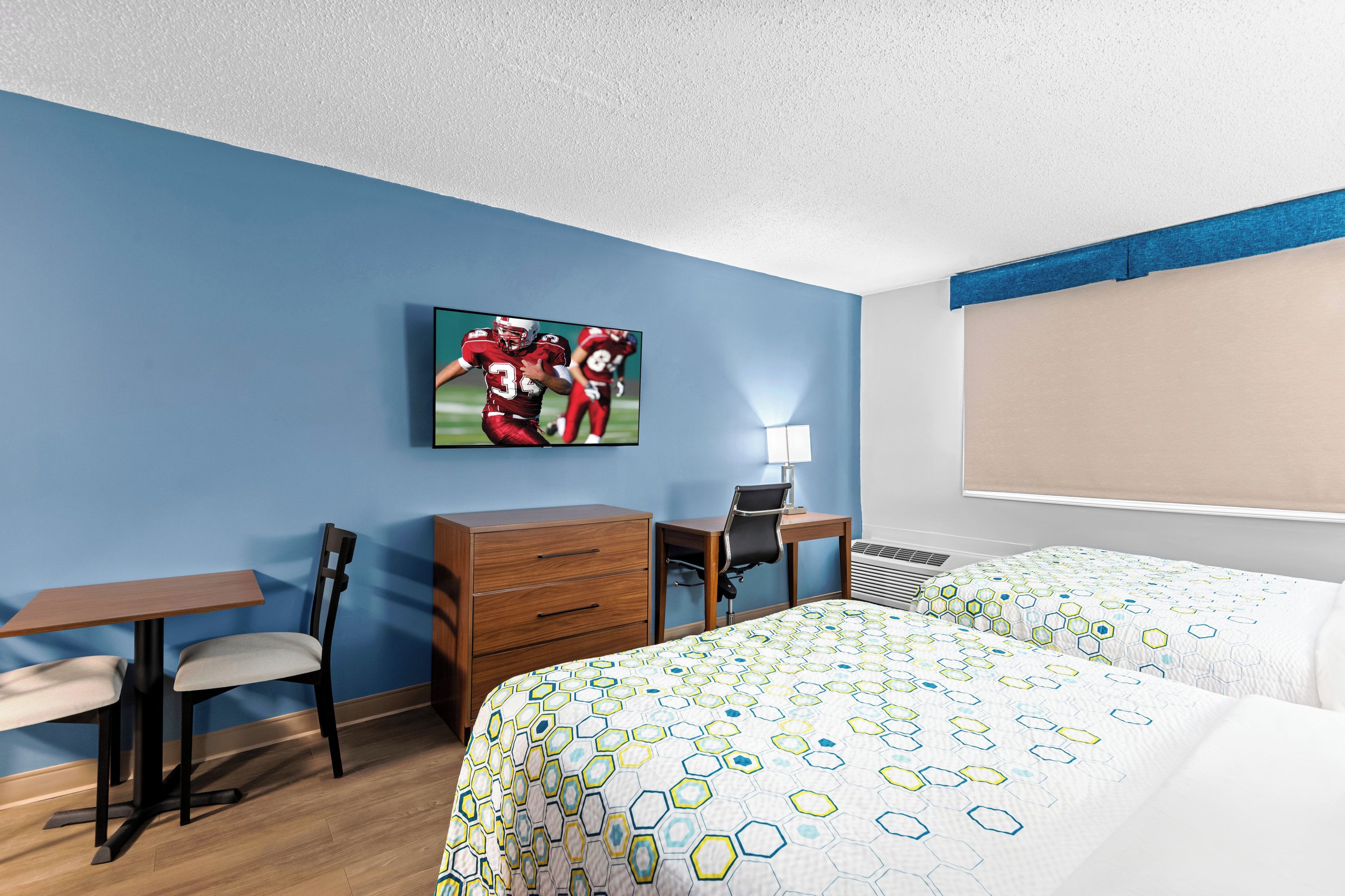 A hotel room with a blue wall, a flat-screen TV, a desk with a chair, a dresser, two beds with patterned bedding, and a window with a beige blind.