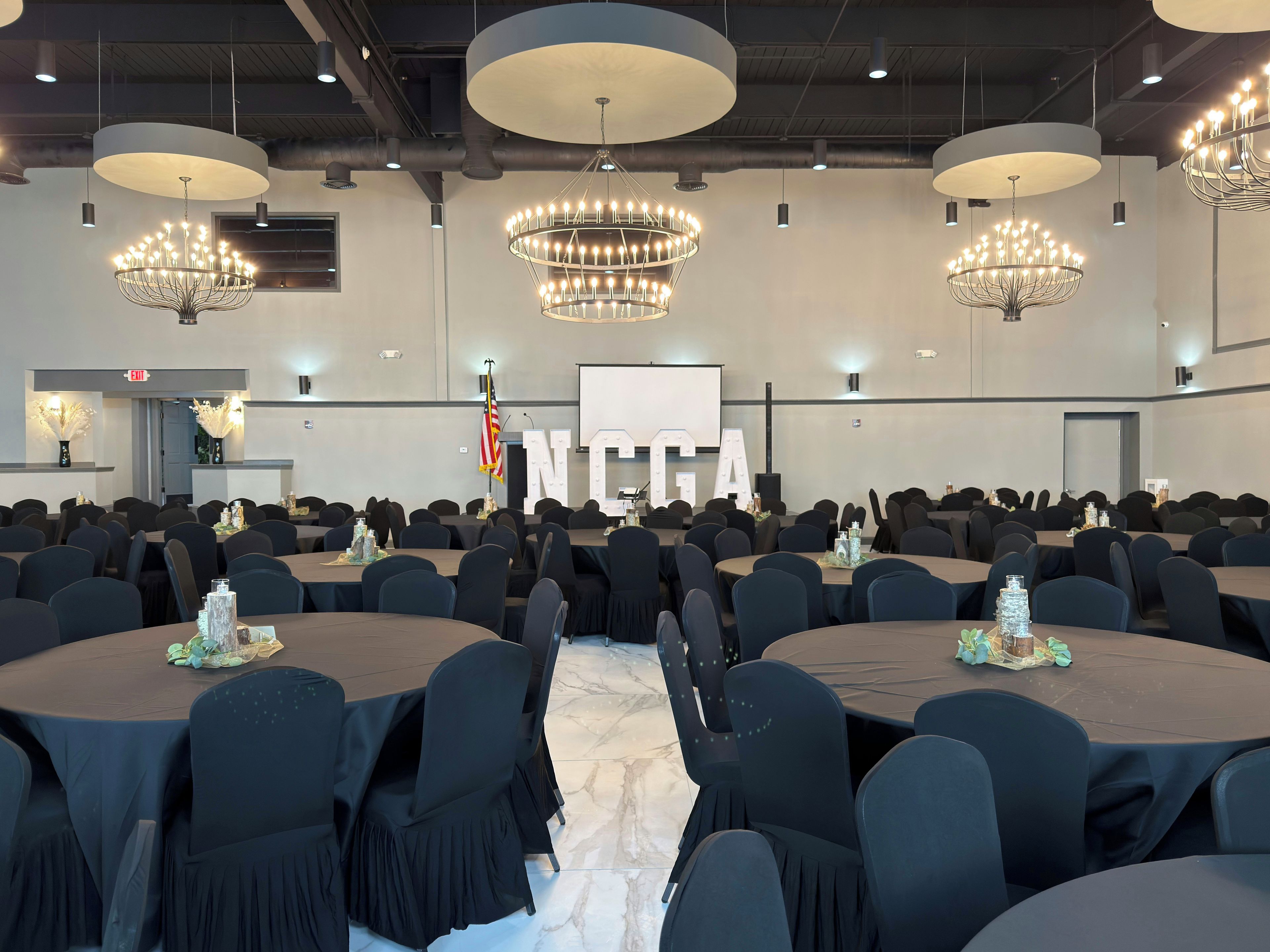 A conference room with round tables, black chairs, chandeliers, and a large screen.