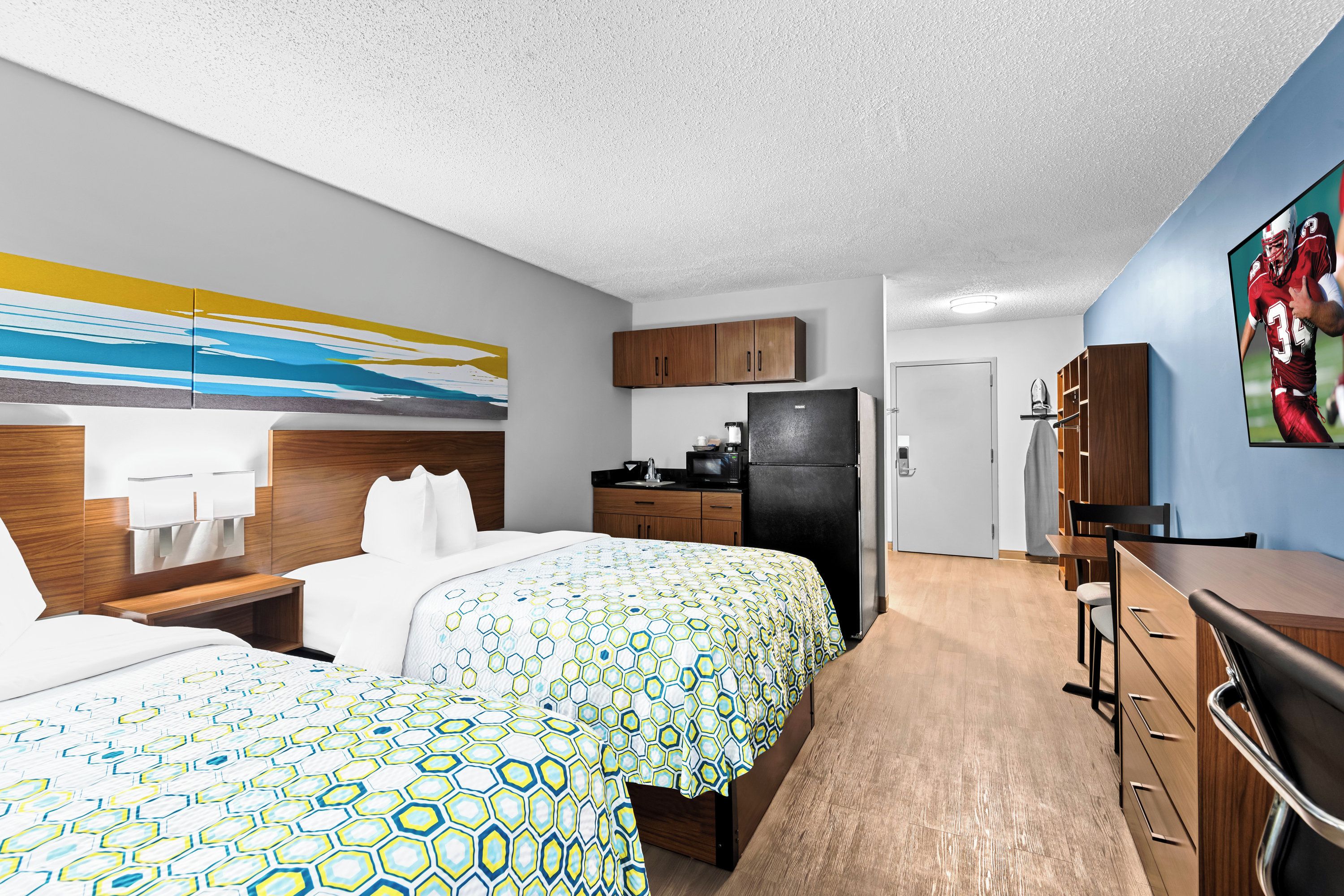 A hotel room with two beds, a kitchenette, a TV, and a painting on the wall.