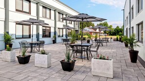 An outdoor patio with tables and chairs, umbrellas, and potted plants. - HomeTowne Studios by Red Roof & Conference Center Cortland (Cortland)