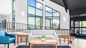 A modern lobby with a grey sofa, a blue armchair, a wooden coffee table, and a staircase with a black railing. - HomeTowne Studios by Red Roof & Conference Center Cortland (Cortland)