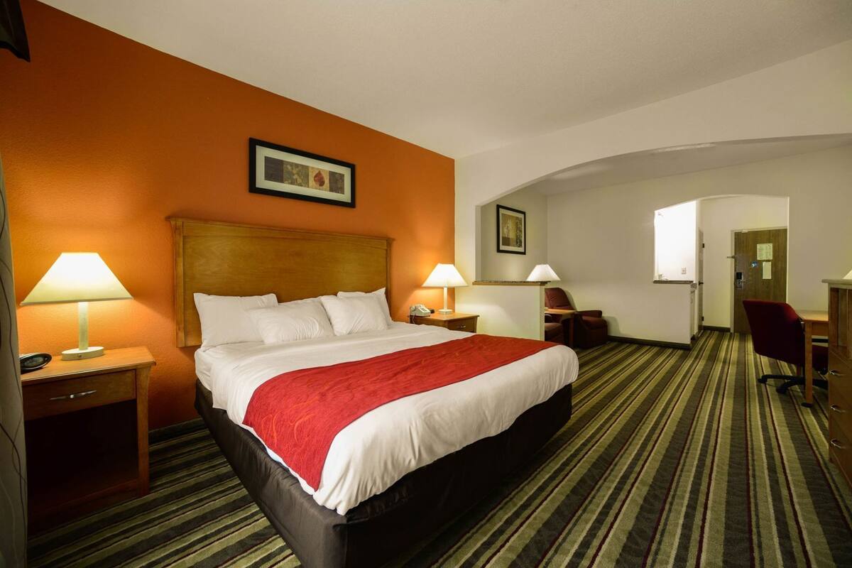 Een hotelkamer met een groot bed, twee nachtkastjes met lampen, een bureau met een stoel, en een deur die naar een andere kamer leidt.