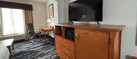 Een hotelkamer met een grote flatscreen televisie aan de muur, een houten kast met lades, een bureau met stoel en een bed met een zwart-wit kleed.