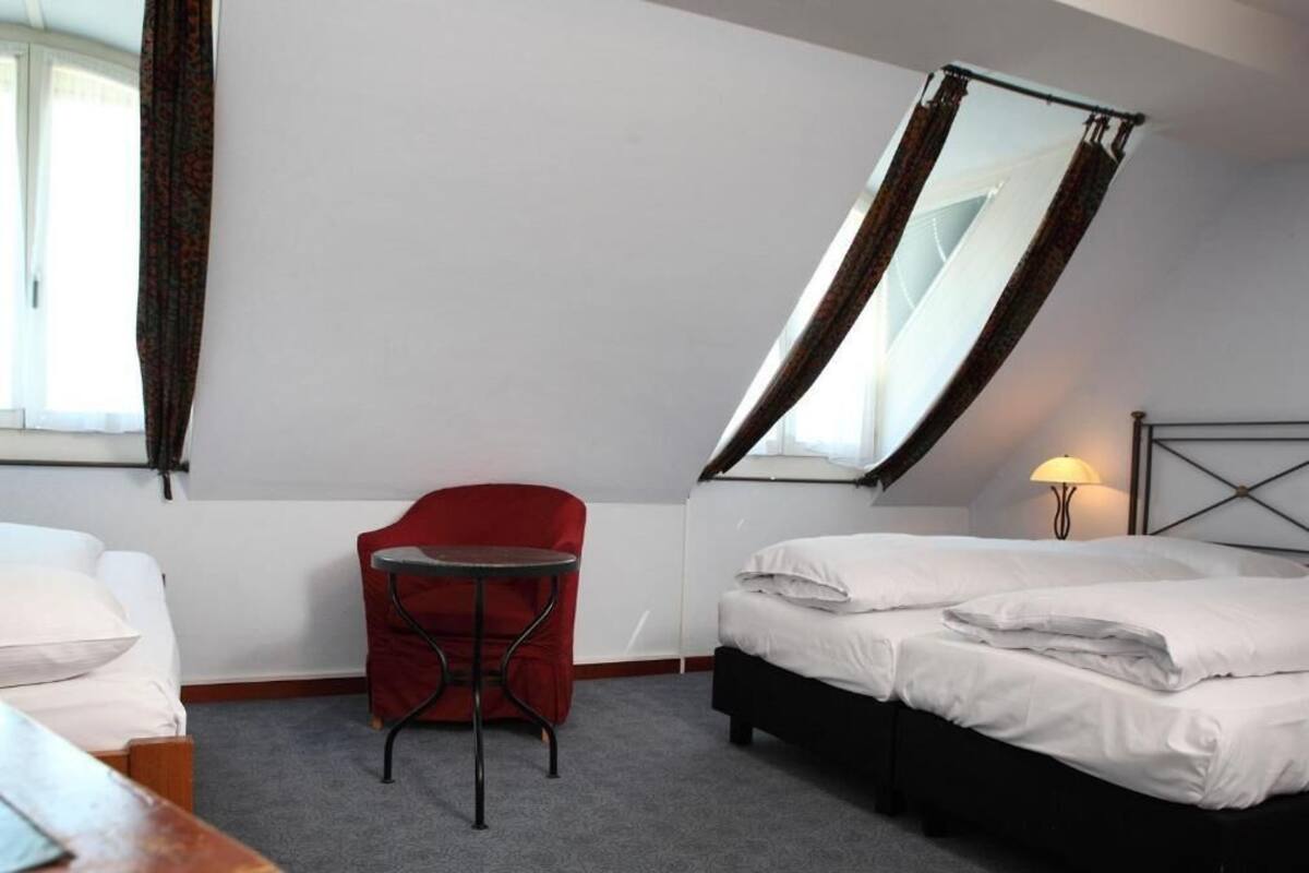 Une chambre d’hôtel avec un lit, une petite chaise rouge, une table et une fenêtre avec des rideaux.