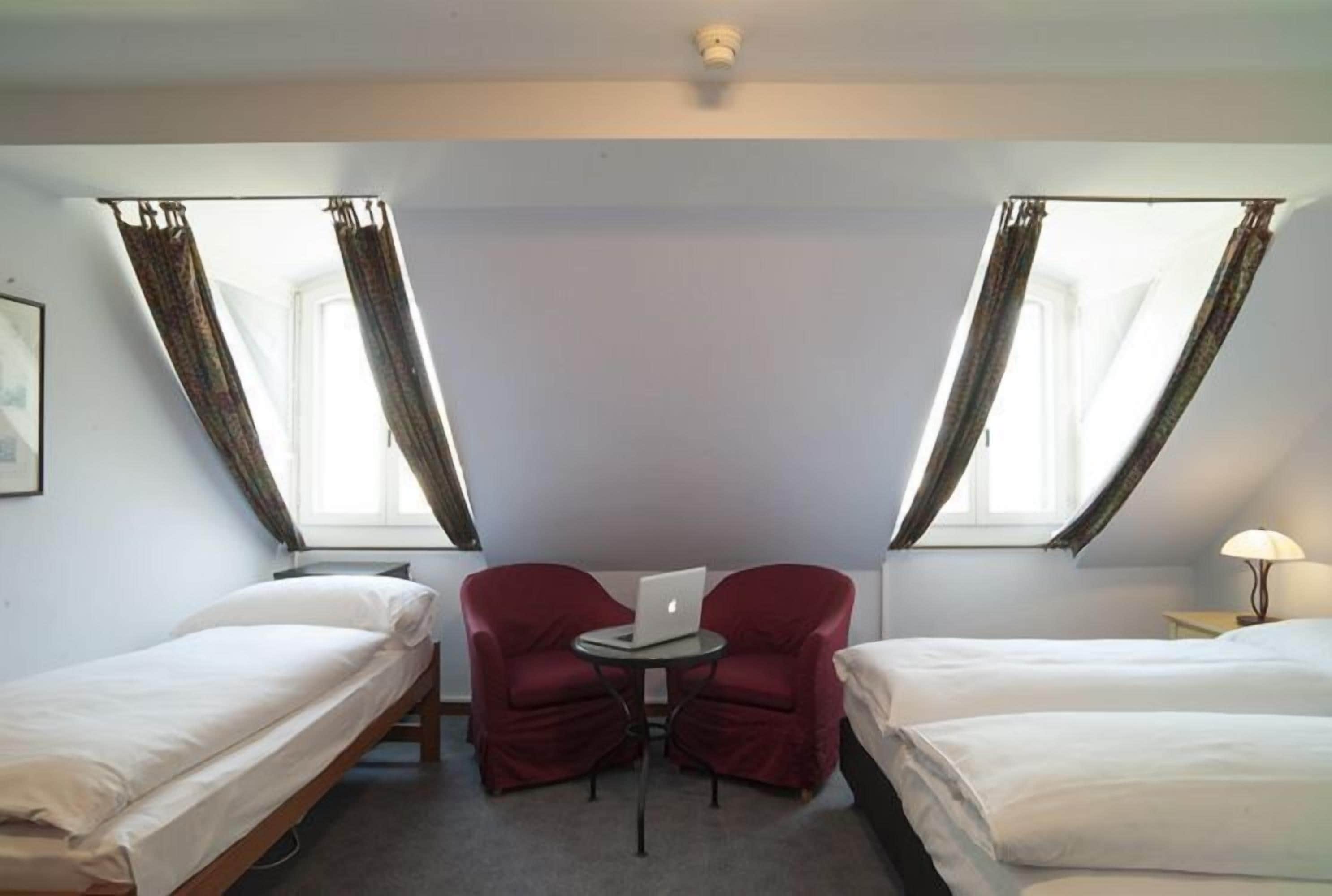 A room with two single beds, a small table with a laptop, two red chairs, and a lamp.