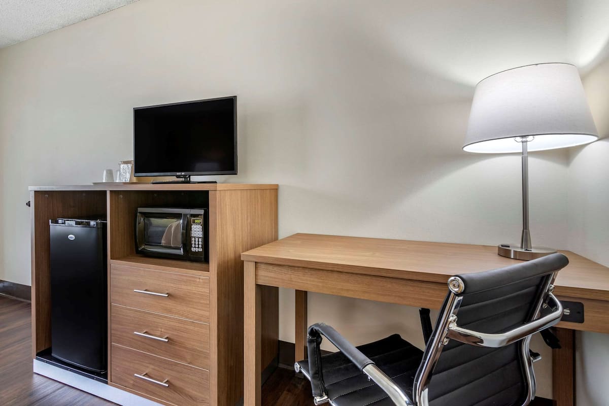 Habitación de hotel compacta con un escritorio de madera, una silla de oficina negra, televisor, microondas y refrigerador.