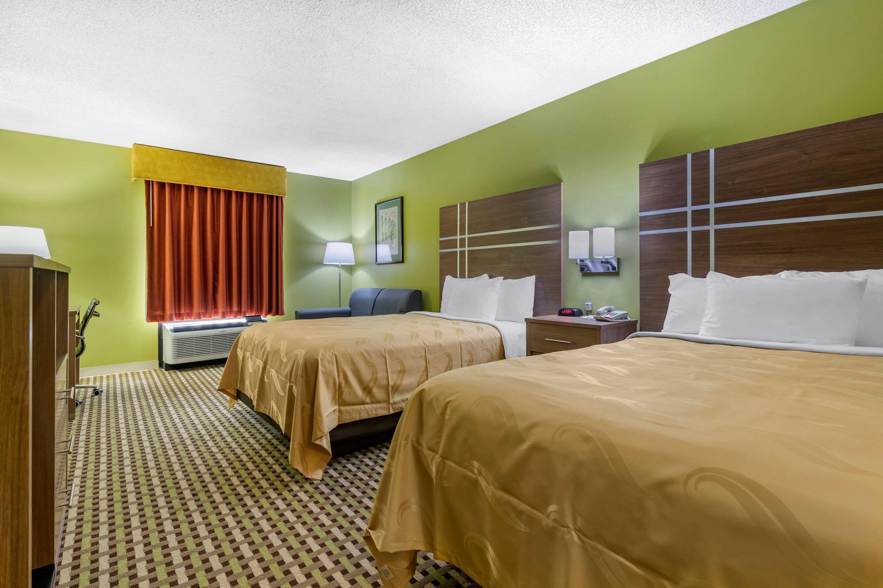 Una habitación de hotel con dos camas, un escritorio, una silla, una lámpara, un reloj y una ventana con cortinas.