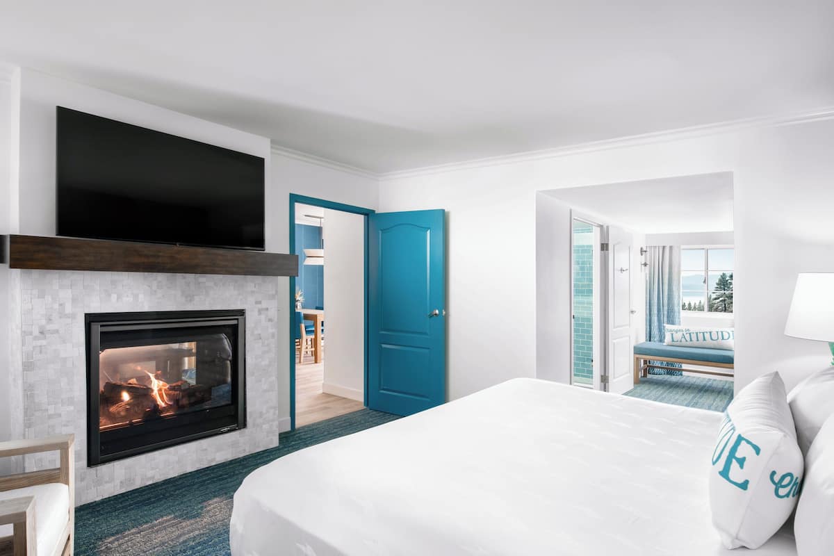 A modern hotel room with a fireplace, a large bed, a blue door, and a balcony with a view.