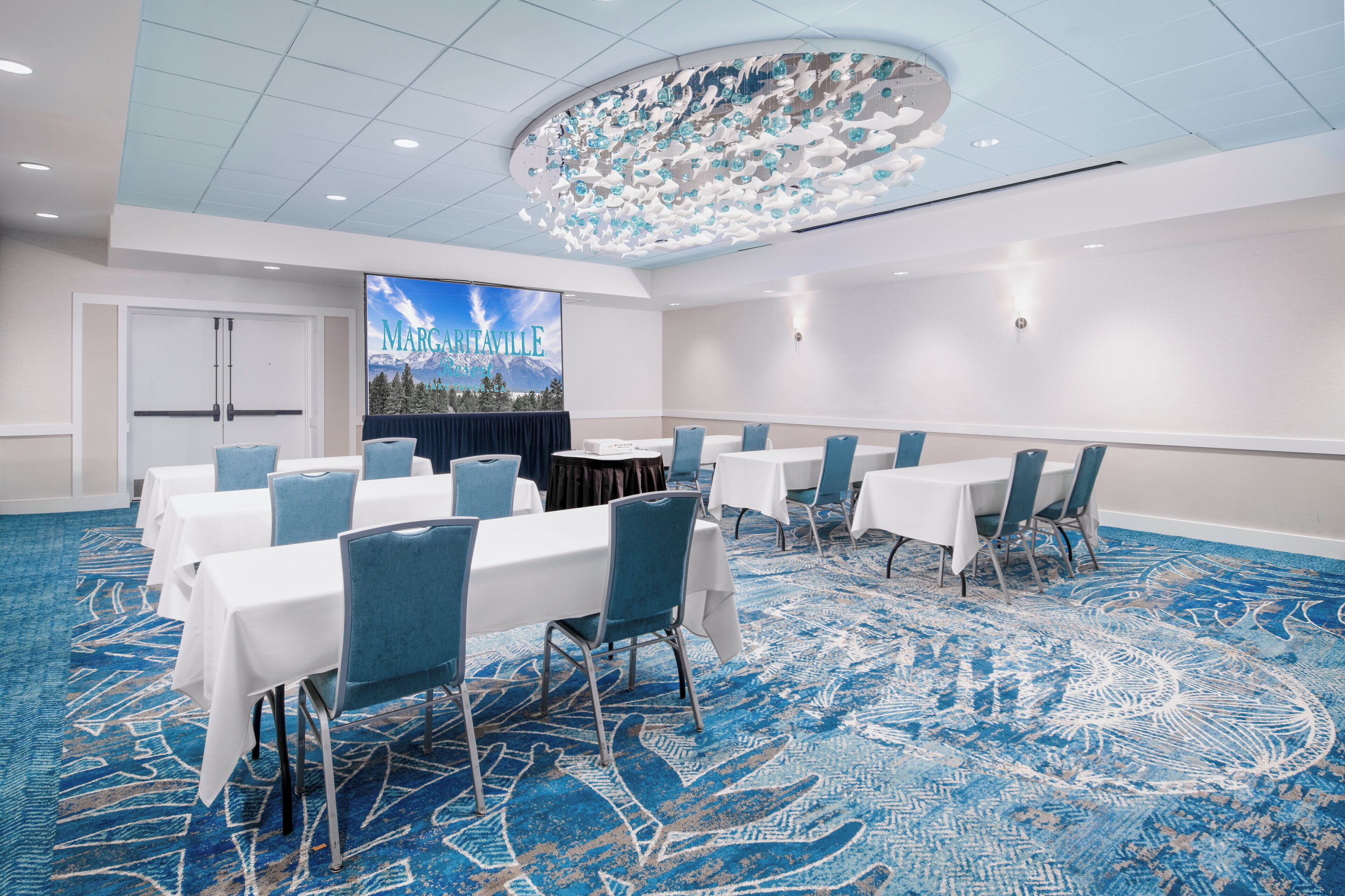 A conference room with white tables and blue chairs, a large screen displaying 'Marcarville', and a patterned carpet.