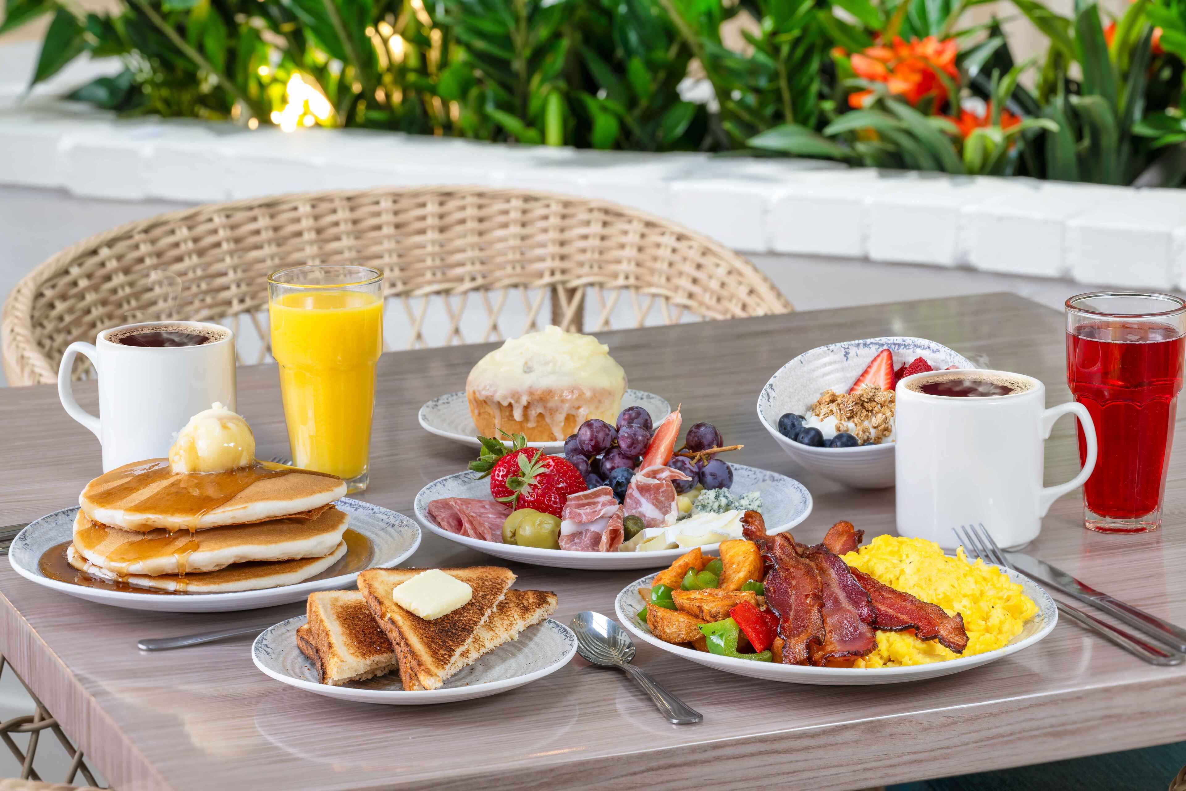 A breakfast table with pancakes, toast, fruit, eggs, bacon, and beverages.