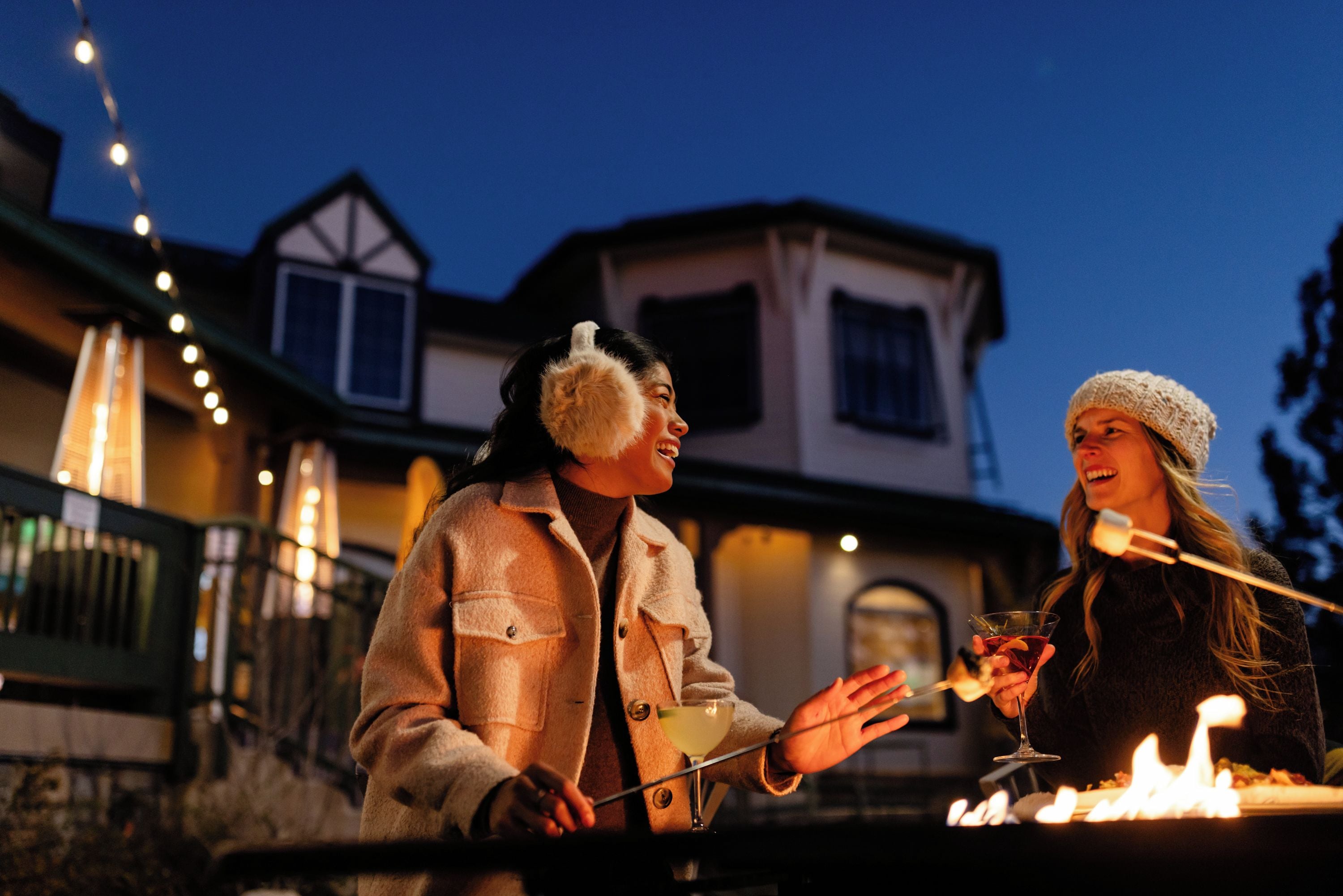 Due persone all'aperto di notte, una con un drink e l'altra con un bastone, con una casa e luci decorative sullo sfondo.