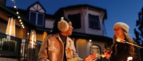 Two people outdoors at night, one holding a drink and the other holding a stick, with a house and string lights in the background.